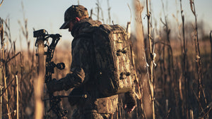 A person in full Veros camouflage outdoor gear stands in a field holding a compound bow, with a large camouflage backpack on their shoulders. The scene is backlit by warm sunlight, creating a strong contrast against the dry reeds.
