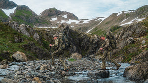 Two hunters backpacking carefully, navigating a rocky stream in a rugged mountain landscape with patches of snow and green vegetation.
