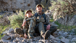 A father and son on a hunt, sitting on a hillside looking up.