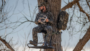 A man in Veros camouflage sits in a tree stand on a tree trunk, with a backpack and additional gear attached to the tree against a bare winter sky.