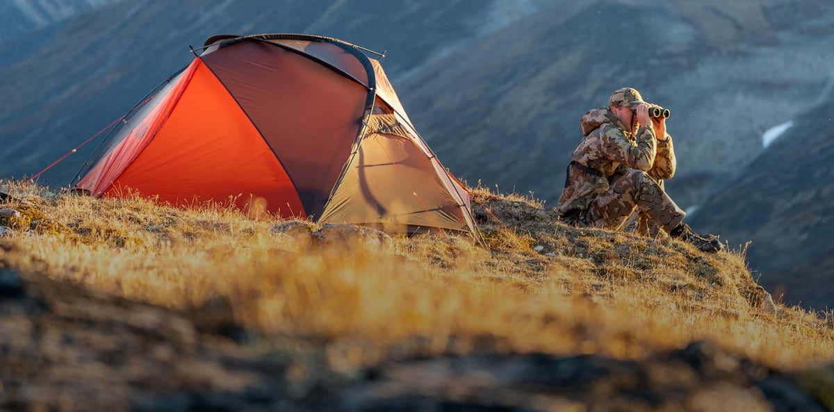Greg McHale in Valo Camouflage looking through binoculars for dall sheep while seated next to orange and gunmetal Mountain Star 2 Person tent