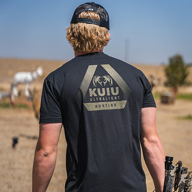 A man wearing KUIU Ultralight Hunting T-Shirt walks in a dry field while carrying a bow.