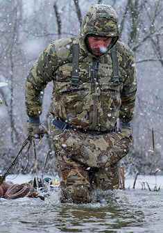 Hunter in a wintry outdoor setting and snow gently falling, wading in shallow water holding multiple duck decoys 