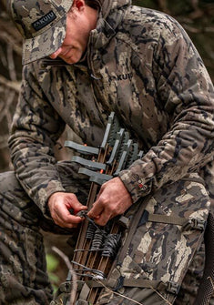 Hunter in-full Veros camouflage hunting jacket, pant and matching cap adjusts a bow loaded with arrows in a wooded setting. 